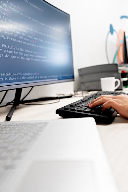A close-up image of a personâ€™s hand operating a black computer keyboard on a white desk, with a large monitor displaying lines of code in the background. To the right of the monitor, there are vario