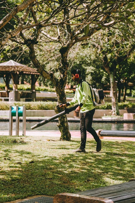 A worker operating a weed trimmer or string trimmer across a paved outdoor area with a surface of dark, weathered bricks, which appear slightly damp. The worker is dressed in a brown apron, sturdy bro