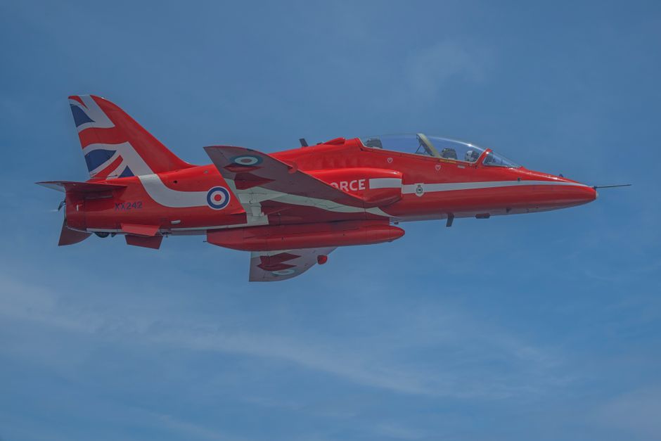 A red military jet aircraft flying in clear blue sky, displaying the UK Royal Air Force roundel on the side and camouflage insignia on the tail fin, with a sleek, aerodynamic structure, sharp nose, an