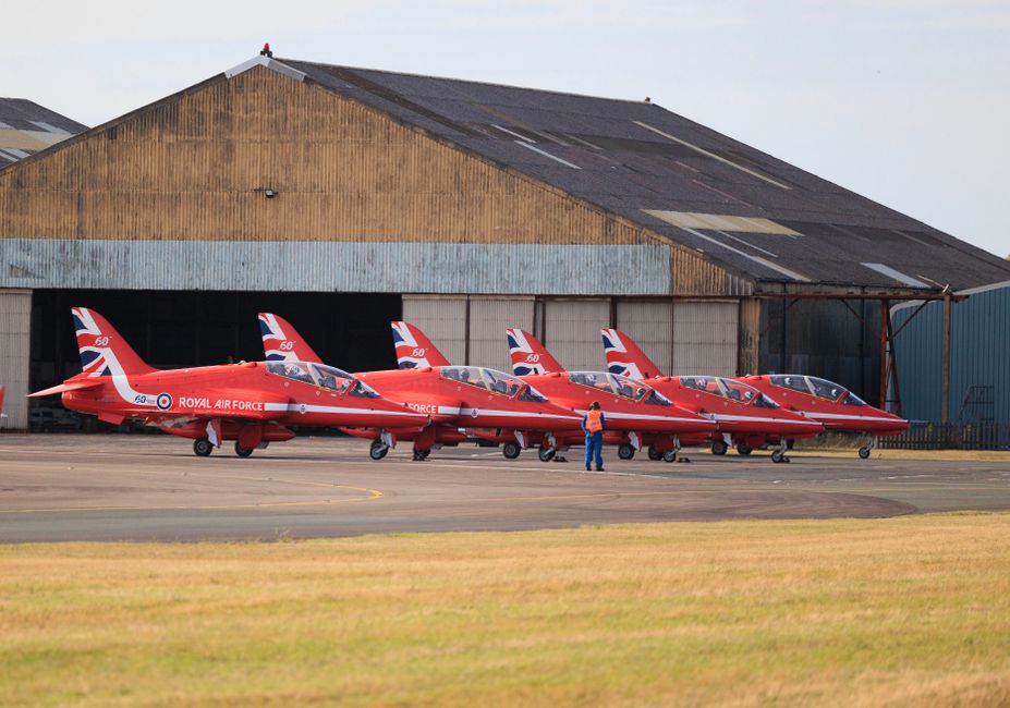 A red military jet aircraft flying in clear blue sky, displaying the UK Royal Air Force roundel on the side and camouflage insignia on the tail fin, with a sleek, aerodynamic structure, sharp nose, an